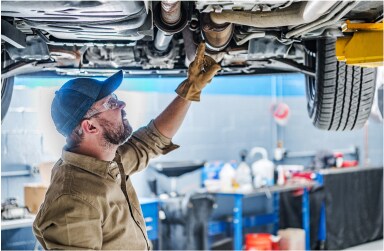 A mechanic is under a lifted car in a garage focusing on the location of the catalytic converter.”