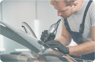 A windshield repair technician wearing overalls and gloves using a tool to repair a car windshield in a well-lit area.